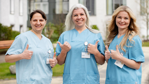 Miteinander Drei Pflegefachfrauen stehen vor dem Pius-Hospital, lächeln in die Kamera und zeigen "Daumen hoch".