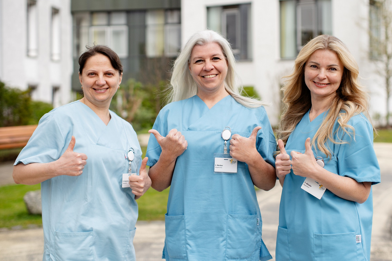 Miteinander Drei Pflegefachfrauen stehen vor dem Pius-Hospital, lächeln in die Kamera und zeigen "Daumen hoch".