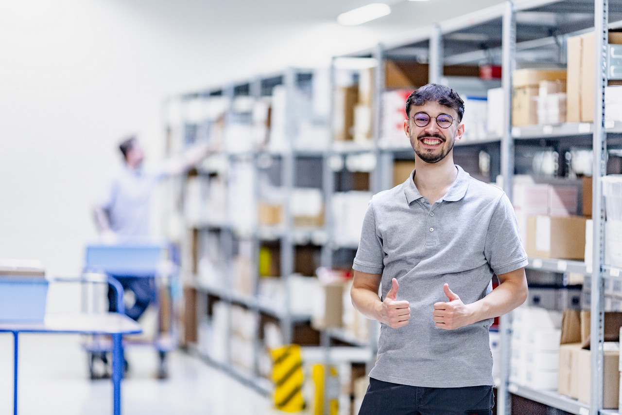 Ein Mitarbeiter der Logistik seht im Lager und blickt lächelnd zur Kamera, beide Daumen hoch