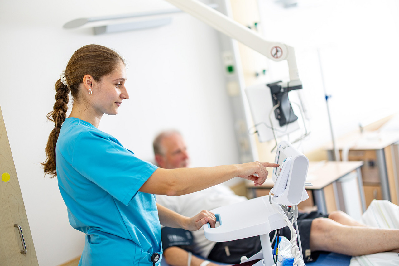 Eine Pflegekraft in blauer Kleidung bedient ein medizinisches Gerät am Bett eines Patienten.