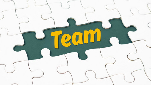 Puzzelbild mit weißen Puzzel-Teilen, in der Mitte ist ein dunkelgrünes Puzzelteil mit dem Wort "Team" in gelber Schrift