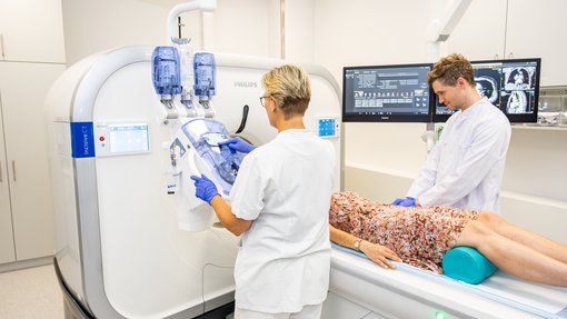 MTA Radiologie Eine MTA bedient ein CT. Im Hintergrund liegt eine Patientin auf dem Tisch des CT und wird von einem Arzt betreut.